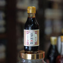 A small bottle of dark soy sauce with a gold cap and cream label sits balanced on the gold lid of a large clear glass jar, with a blurred background of other bottles.