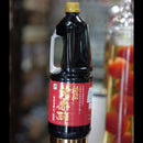 A large plastic bottle of dark soy sauce with a red label and gold lettering, topped with a yellow cap, set against a blurred background of a shop interior.