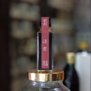 A small rectangular bottle with a red label and gold cap, sealed inside a large clear glass jar with a gold lid, photographed against a blurred dark background.