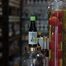 A small dark bottle with a green cap and a white label featuring black Japanese characters, placed on top of a large clear glass jar filled with brown beans. In the background, a tall glass jar holds red fruits in yellow liquid, set in a softly blurred shop environment.